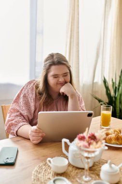 Sıcak bir sabah sahnesinde Down sendromlu genç bir kadın tabletinde kahvaltı ederken görülüyor..