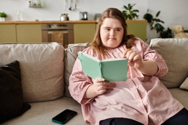 Down sendromlu genç ve güzel bir kadın rahat bir kanepede rahatlar, odaklanarak ve neşeyle kitap okur..