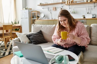 Rahat yerinde, genç bir kadın dizüstü bilgisayarını kullanırken sıcak bir içkinin keyfini çıkarıyor..