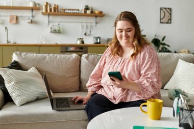 Down sendromlu genç bir kadın kanepede oturuyor, yanında dizüstü bilgisayarıyla akıllı telefonunu kullanıyor..