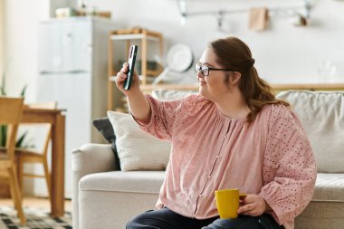 Down sendromlu genç bir kadın evde elinde telefon ve kahveyle gülümsüyor..