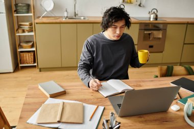 A handsome young Asian man sits at his kitchen table, focused on studying online, enjoying a drink. clipart