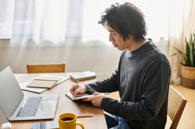 Genç bir adam özenle evinden ders çalışıyor, bilgisayarına odaklanırken notlar alıyor..