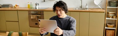 Çağdaş mutfağında online çalışan genç bir adam tablet ekranına, afişine dikkatle odaklanıyor.