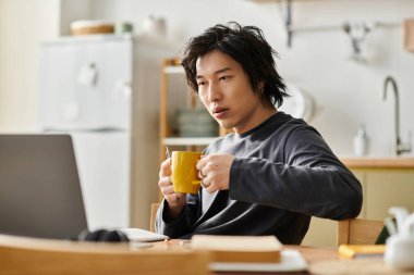 Yakışıklı bir Asyalı genç öğrenci sıcak bir içkinin tadını çıkarırken online çalışmalarına konsantre oluyor..