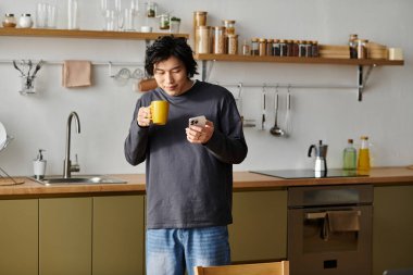 Genç Asyalı bir adam şık mutfağında duruyor, içkisini yudumluyor ve telefonunu kontrol ediyor..