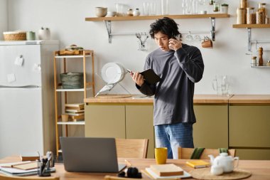 Genç bir Asyalı ev mutfağından internetten ders çalışıyor. Defter ve telefonla birden fazla iş yapıyor..