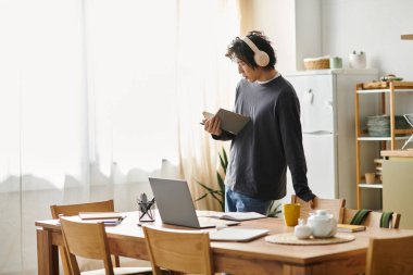 Yakışıklı genç bir adam, elinde bir kitap ve dizüstü bilgisayarla evde internetten çalışıyor..