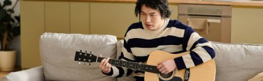 Yakışıklı genç Asyalı bir adam, akustik gitar ve pankart çalmayı öğrenirken koltuğa oturur.