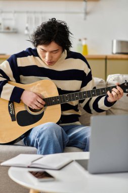 Genç Asyalı bir adam akustik gitarına dikkatle odaklanıyor, evde melodi çalışıyor..