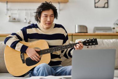 Genç bir Asyalı, parlak bir oturma odasında akustik gitar çalıyor. Dersine odaklanmış..