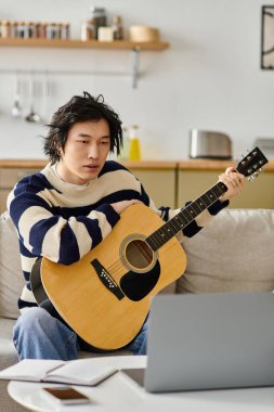 Genç Asyalı adam evde rahat bir ortamda akustik gitar öğrenmeye odaklanmış..