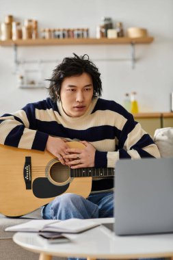 Yakışıklı Asyalı genç adam rahat bir oturma odasında akustik gitar çalıyor, öğrenmeye hevesli..