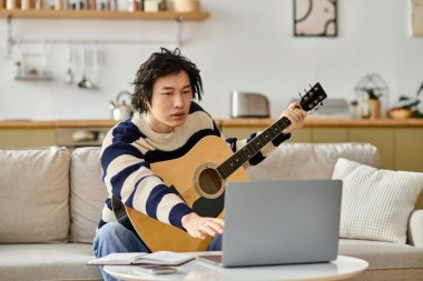 Yakışıklı bir genç adam akustik gitar çalmayı dikkatle öğreniyor, dizüstü bilgisayarına odaklanıyor..