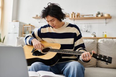 Genç Asyalı bir adam kanepede oturup akustik gitarı bir kitaptan öğrenmeye odaklanmış..
