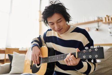 Genç bir adam akustik gitarını çalışıyor, müzik dersine odaklanmış ve dalmış..