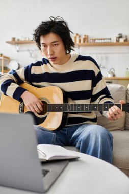 Genç Asyalı bir adam rahat bir oturma odasında akustik gitar çalıyor, öğrenmeye odaklanmış..
