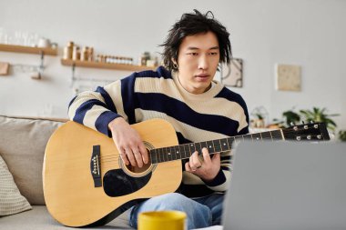 Genç Asyalı bir adam koltukta rahatça oturur akustik gitar çalmayı öğrenir, odaklanır ve ilham verir..