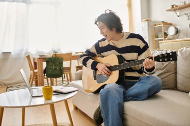Yakışıklı genç adam evinde rahatça otururken akustik gitar çalma pratiği yapıyor..