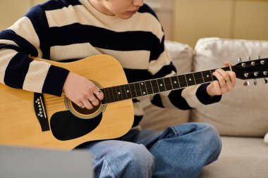 Çizgili kazak giymiş genç bir adam kanepede akustik gitar çalıyor..