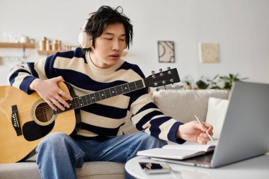 Yakışıklı Asyalı genç adam şık bir ev ortamında notlar alırken akustik gitar çalıyor..