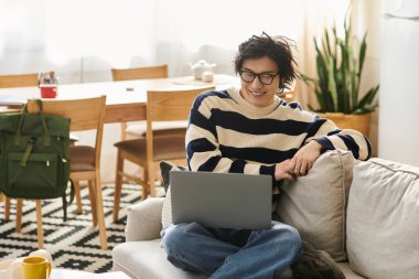Yakışıklı Asyalı genç adam evinin rahat bir köşesinden laptopuyla çalışırken gülümsüyor..
