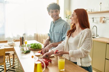 Genç bir çift, parlak mutfaklarında birlikte yemek pişirirken gülüşlerini ve kahkahalarını paylaşıyor..