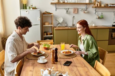 Genç bir çift, mutfak masasında ev yapımı yemek yerken kahkaha ve neşeyi paylaşıyor..