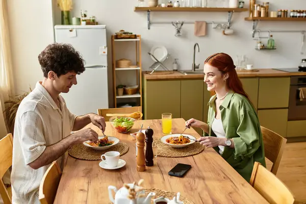 Genç bir çift, mutfak masasında ev yapımı yemek yerken kahkaha ve neşeyi paylaşıyor..