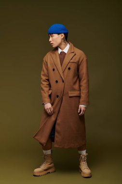 Modaya uygun bir ceket ve mavi bir bere giyen Asyalı genç bir adam stüdyo ortamında gösterişli bir şekilde duruyor..