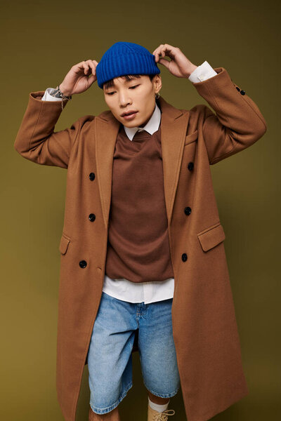 A young man flaunts a stylish brown coat and blue beanie in a modern backdrop.