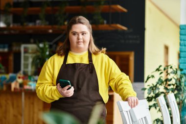 Down sendromlu genç bir kadın bir kafede gelişigüzel giyinmiş elinde kendine güveni olan bir telefon tutuyor..