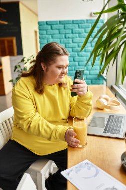 Down sendromlu genç bir kadın bir kafede oturmuş smoothie yudumluyor ve telefonunu kullanıyor..