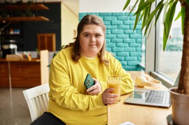 Down sendromlu genç bir kadın bir kafede oturmuş, telefonunu kullanırken elinde içkisini tutuyor..