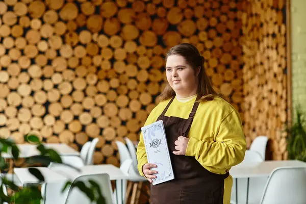 Down sendromlu genç bir kadın bir kafede elinde menüyle gururla duruyor ve sıcaklığını paylaşıyor..