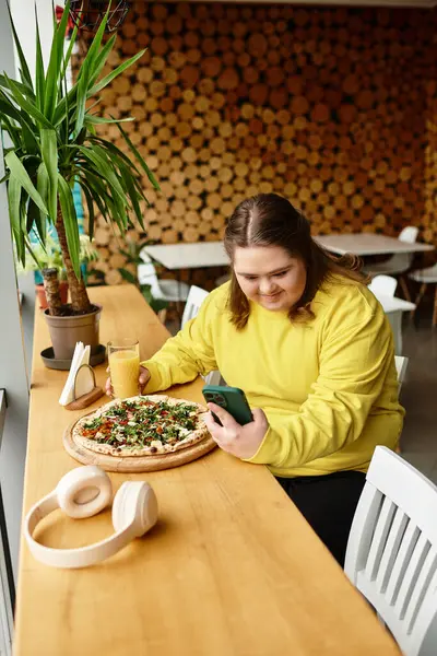 Down sendromlu genç bir kadın telefonuna gülümserken kafede pizza yiyip meyve suyu içiyor..