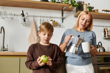 Bir kadın, oğlu elmayı tutarken kahve yapar. Mutfağın sıcacık rutinini vurgular..