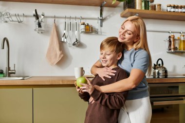 Annesi onu mutfakta nazikçe kucaklarken bir çocuk mutlu bir şekilde yeşil bir elma tutuyor..