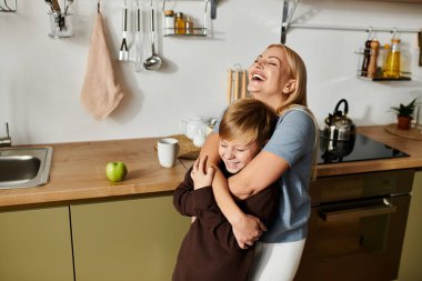 Çağdaş bir mutfakta gülümseyen oğlunu kucaklayan bir kadın neşe ve bağlantı yayar..