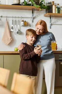 Sıcak bir mutfakta, bir anne ve oğlu birlikte akıllı telefona bakarken gülümserler..