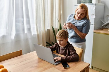 Bir kadın sıcak bir sohbeti paylaşırken mutfak masasında oğluna ödevlerinde yardım eder..