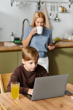 Oğlu mutfak masasında dizüstü bilgisayarına odaklanırken bir kadın sıcak bir içkiden zevk alır..