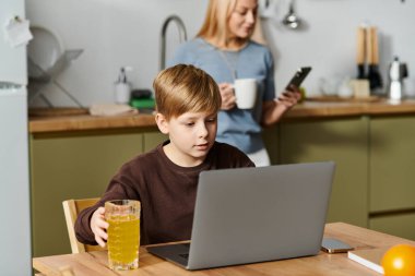Annesi sıcak bir içkinin keyfini çıkarırken bir çocuk dizüstü bilgisayarla etkileşime giriyor ve telefonunu yakınlarda kontrol ediyor..