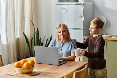 Bir kadın dizüstü bilgisayarında çalışırken oğlu ona sıcak bir içki ikram ediyor ve güçlü bağlarını gösteriyor..