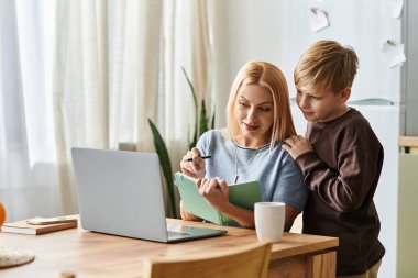 Bir anne, güneşli bir öğleden sonranın tadını çıkarırken oğluna tahtadan bir masada ödevlerinde yardım eder..