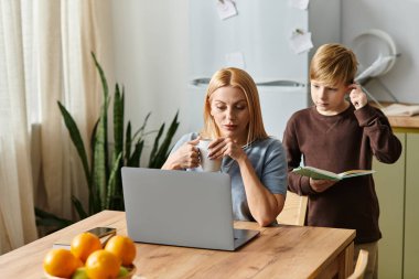 Bir anne oğluna mutfak masasında bir kitapla yardım ederken sıcak bir içkiden zevk alır..