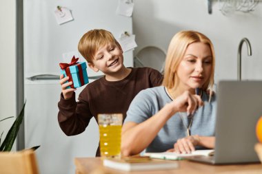 Neşeli bir çocuk, annesi dizüstü bilgisayarında aile sevgisini gösterirken hediye verir..