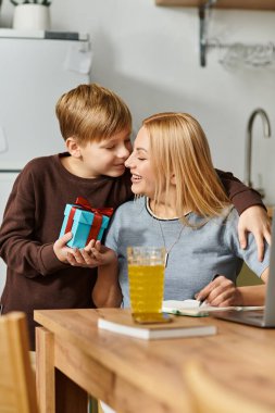 Neşeli bir anne ve oğlu bir hediyeyle mutfak masasında sevgi dolu bir anı paylaşıyorlar..