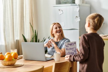Bir oğul, annesine el yapımı bir kartla sürpriz yapar. O da evinde dizüstü bilgisayarda çalışır..