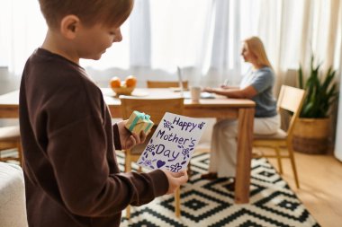Neşeli bir çocuk, annesi yemek masasında çalışırken bir el yapımı kart tutuyor..